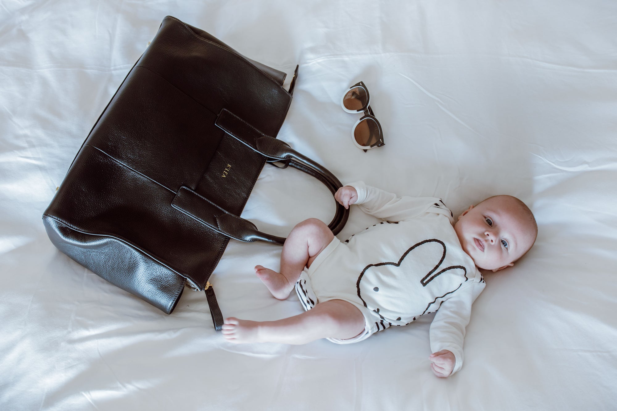 Baby lying on a white surface next to a Belle black Diaper handbag