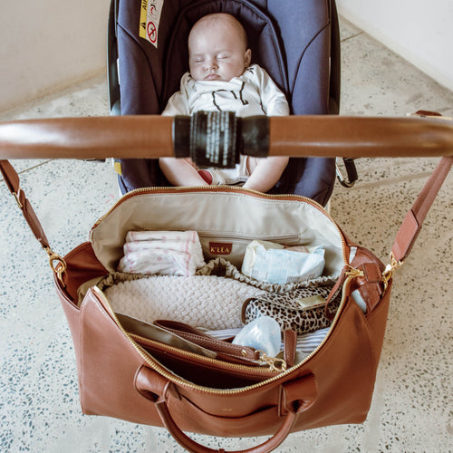 Tan genuine leather bag open with baby items, including a baby in a stroller in the background.